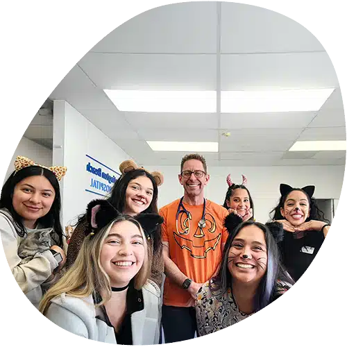 A group of smiling people pose indoors, many wearing animal ear headbands and face paint. One veterinarian at the center sports an orange shirt with a jack-o’-lantern design. The festive, cheerful atmosphere hints at a vet-themed celebration.