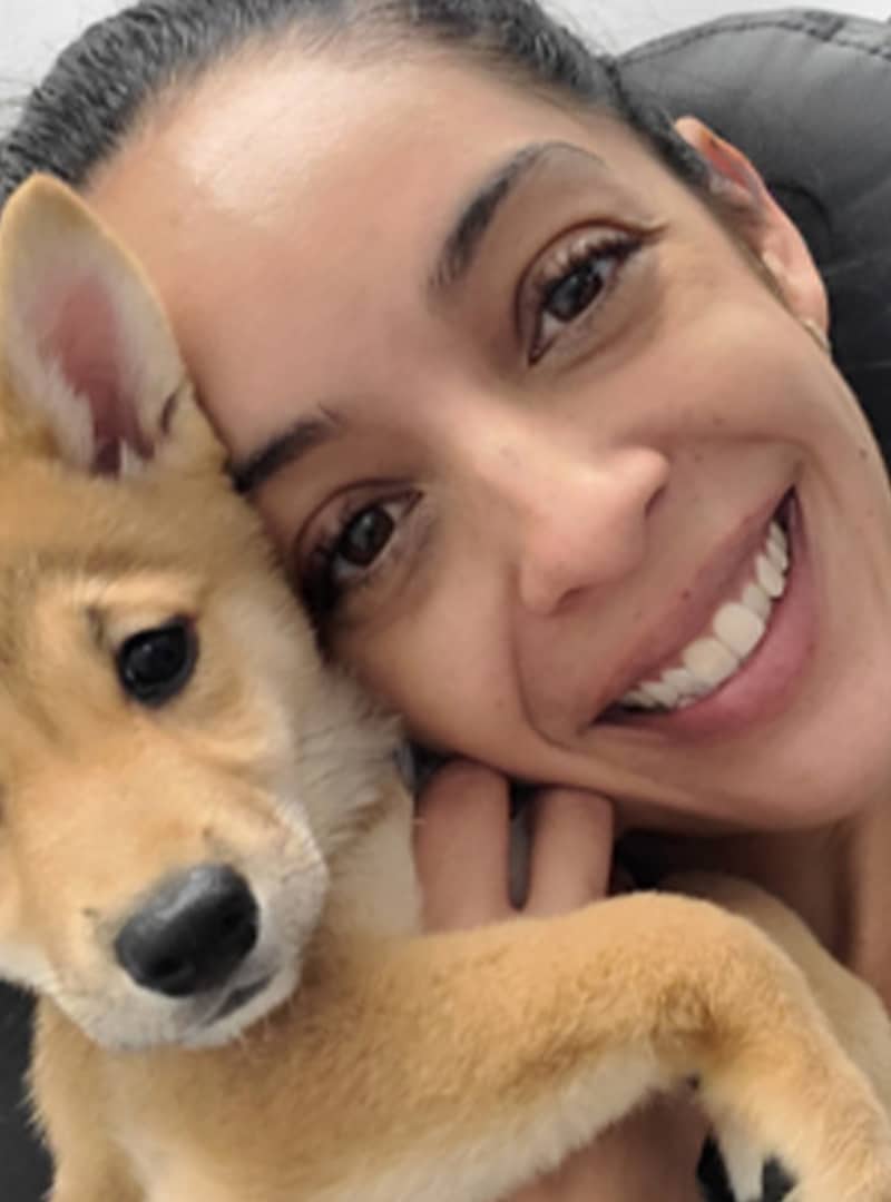 A woman, possibly a veterinarian, smiles closely next to a tan and white puppy with pointy ears, both looking at the camera.